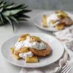Two white plates of Grilled Pineapple Shortcake topped with whipped cream and next to the top of a pineapple.