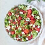 White shallow bowl filled with diced Watermelon, peeled cucumber ribbons, sliced basil, goat cheese, almonds, and lime.