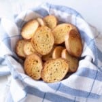 Golden brown oven baked crostini in a blue and white striped towel lined basket.