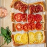 A sliced heirloom tomato puff pastry tart with whipped goat cheese on a cutting board next to basil and a glass of rosé wine.