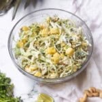 A clear bowl of pineapple coleslaw next to cilantro, lime wedges, cashews, and the top of a pineapple.