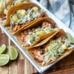 A platter of salmon tacos with pineapple slaw next to lime wedges and a basket of corn tortillas.