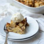 Slice of caramelized apple bread pudding topped with a scoop of vanilla ice cream on a white plate with a fork.