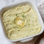 Crème Fraîche Mashed Potatoes topped with chives and butter in a white baking dish next to a wooden spoon and white towel.