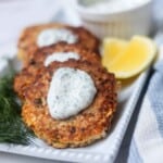 Salmon patties with dill sauce on a white platter next to lemon wedges, fresh dill, and extra sauce.
