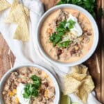 Two bowls of white ground chicken chili topped with sour cream, cilantro, and cheese on a wooden board next to chips and cilantro.