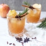 Two sparkling apple cider mocktails in a clear glass garnished with cinnamon, spices, rosemary, and apple.