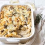 Baked mushroom mac and cheese in a white baking dish with a scoop missing next to a sprig of thyme and white wine.