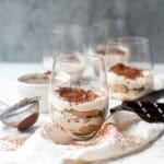 A clear glass of a mini tiramisu cup on top of a cream towel and next to a sifter with cocoa powder in it.