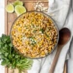 Mexican street corn elote pasta salad in a glass bowl next to cilantro, lime wedges, cotija, and a wooden spoon.